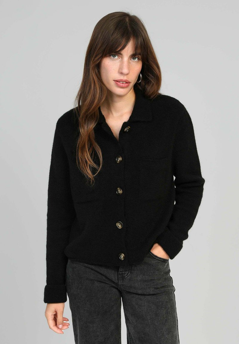 Cardigan noir à boutons fabriqué dans un matériau doux, avec un col, deux poches avant et de grands boutons marron. Manches longues avec détail au niveau des poignets.