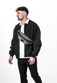 Bolso cruzado gris fabricado con un material de textura suave y detalles en negro. Presenta una forma curva y una correa ajustable.