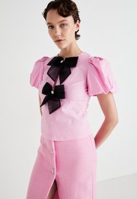 Kvinne som har på seg en rosa topp med puffermer, to store svarte sløyfer og et høy midje rosa skjørt med splitt foran og knappdetaljer.