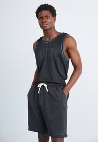 Svart ermeløs trenings tank topp med teal trim, med teksten "Fleisse". Matchet med mørkegrå shorts og hvit snøring.