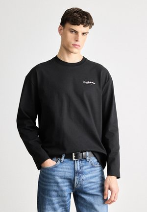 Tricou negru cu mânecă lungă, din bumbac, cu decolteu rotund și un mic logo alb pe piept, asortat cu blugi rupți de culoare albastră și curea neagră.