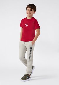 Gutt som står med hendene i lommene, iført en rød Champion t-skjorte, lysegrå joggebukser med logo, og grønne sneakers med hvite såler.
