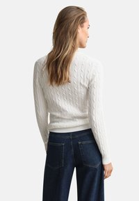 Weißer Strickpullover mit Kabelmuster, geripptem Saum und Bündchen, kombiniert mit dunkelblauen Jeans mit Gesäßtaschen und geradem Bein.