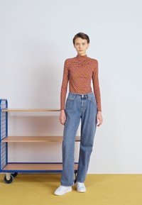 Top a maniche lunghe marrone con un motivo geometrico colorato, abbinato a jeans in denim chiaro a vita alta e sneakers bianche. Sfondo semplice.