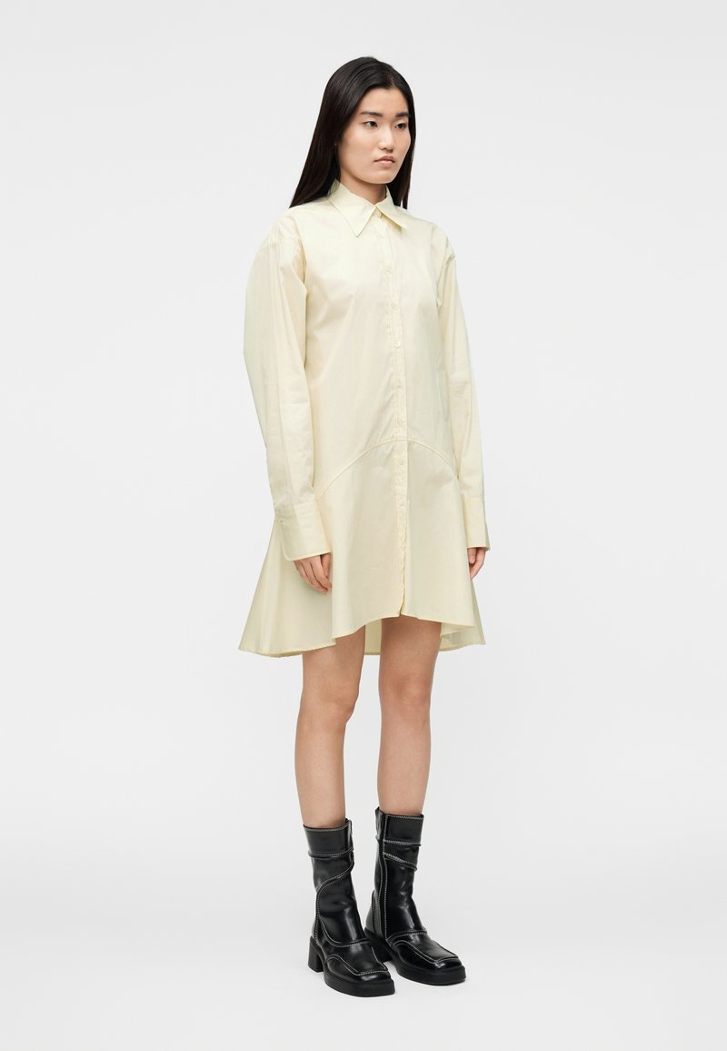 Robe chemise jaune pâle à manches longues avec fermeture à boutons, col et ourlet évasé. Associée à des bottes de combat noires montant jusqu'aux genoux.