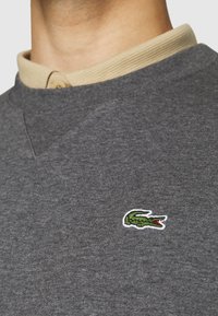 Šedý bavlněný svetr s texturovaným povrchem, který má béžový žebrovaný límec a na hrudi je vyšitý malý zelený krokodýl logo.