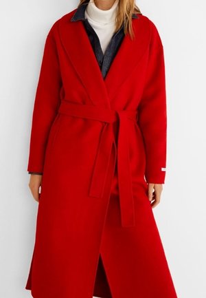 Woman wearing a long red belted coat over a dark denim shirt and white turtleneck sweater, standing against a white background.