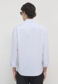 Homme portant une chemise blanche à manches longues avec de fines rayures verticales et un pantalon noir, de dos avec des manches retroussées et des lunettes de soleil noires.