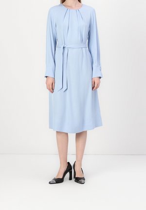 Mujer que lleva un vestido midi de manga larga de color azul claro con un lazo en la cintura, combinado con zapatos negros de tacón alto con punteras metálicas.