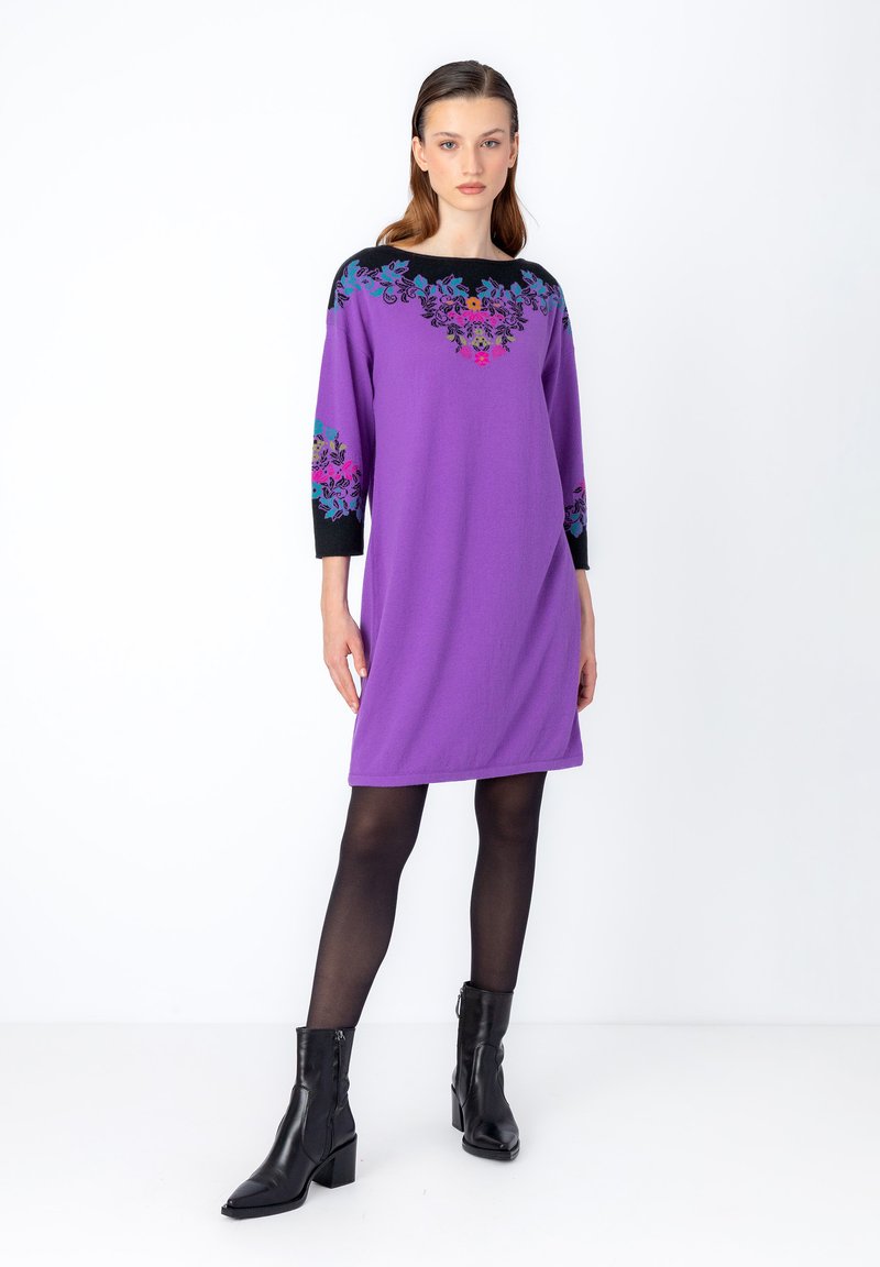 Violetti mekko mustilla yksityiskohdilla ja kukkakirjonnalla hihoissa. Valmistettu pehmeästä kankaasta, rento malli ja pyöreä kaula-aukko. Mustat nilkkasaappaat.