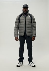 Grå pufferjakke med glidelås foran, svarte bukser og hvite sneakers. Solid tekstur, polstret design, med en ryggsekk og caps.