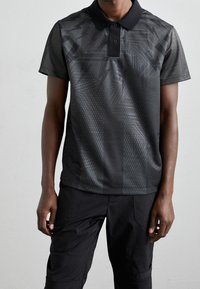 Černé polo tričko s texturovanými geometrickými vzory, krátkými rukávy a klasickým límcem se třemi knoflíky. Nosené s černými kalhotami.