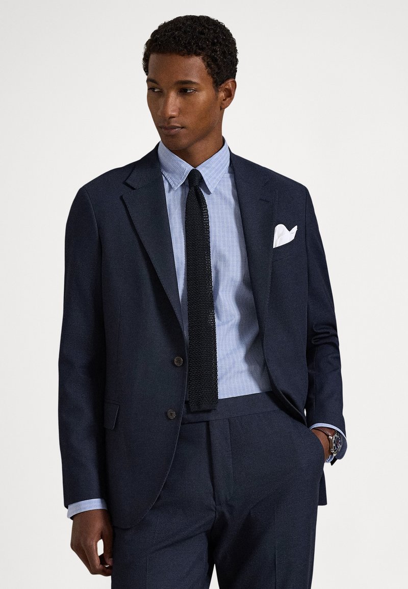 Vīrietis tumsnēji zils kostīmā ar gaiši zilu rūtainu kreklu, melnu adītu kaklasaiti, baltu kabatas lakatiņu un pulksteni, pozējot ar vienu roku kabatā.