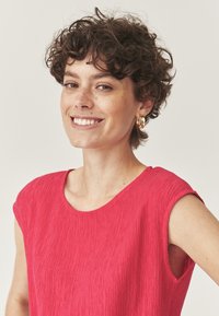 Jasnoružová textúrovaná blúzka s okrúhlym výstrihom a krátkymi rukávmi. Tkanina má jemný, zvrásnený vzor. Viditeľné sú aj kruhové zlaté náušnice.