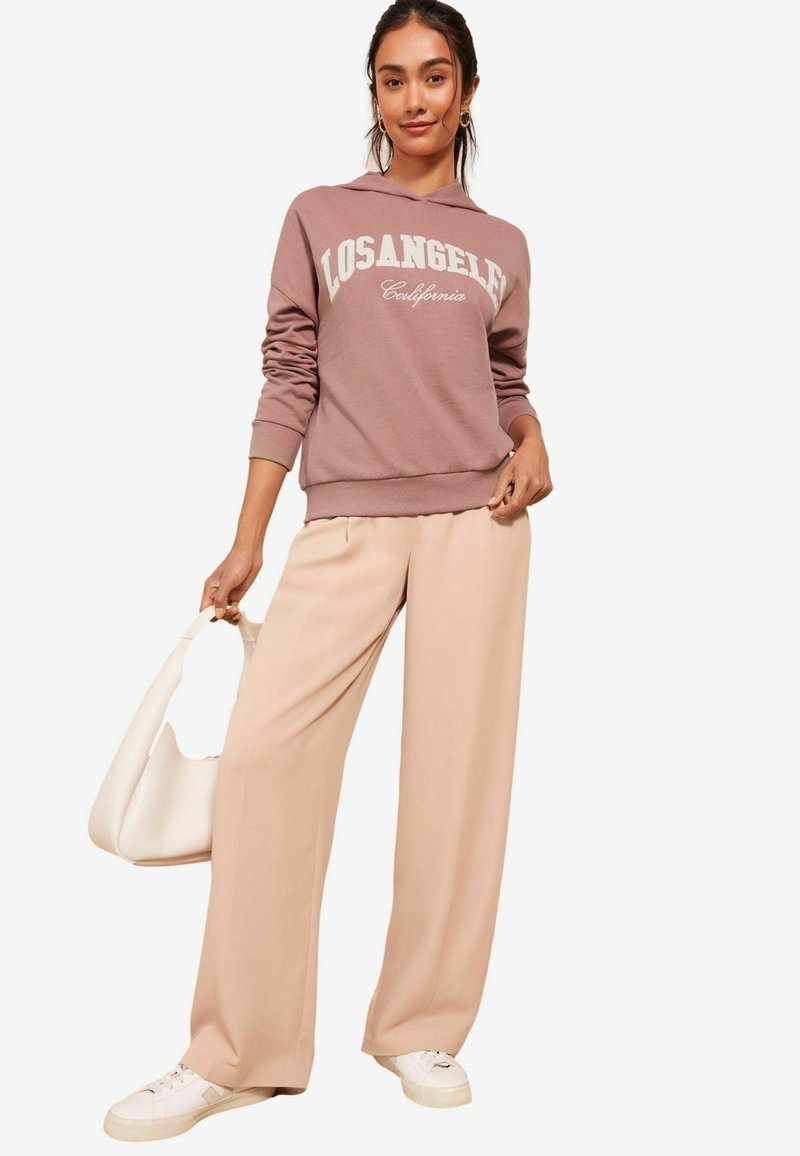 Sudadera de color rosa polvo con el texto "LOSANGELES California", combinada con pantalones de pierna ancha beige y zapatillas blancas. Tejido suave, corte casual.