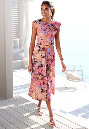 Blumenkleid Maxi mit rosa, orange und lila Farbtönen, kurzen Ärmeln und einem taillierten Gürtel. Getragen mit beigen Sandalen auf einem Holzboden.