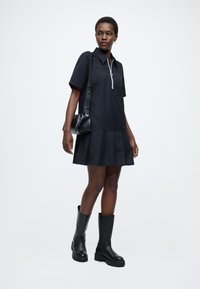 Μαύρο φόρεμα με κοντά μανίκια, γιακά με φερμουάρ, φούστα με φλάρε και πιέτες. Συνδυάζεται με μαύρες μπότες μέχρι το γόνατο και μικρή μαύρη τσάντα.