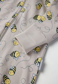 Beige Zip-Hoodie-Einteiler mit einem Muster aus lächelnden Bienen mit gelben Körpern und schwarzen Streifen, akzentuiert durch weiße Flügel und geschwungene Linien.