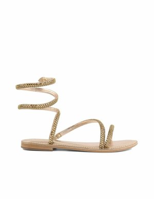 Sandalo beige piatto con cinturini borchiati d’oro, inclusa la fascia per l’alluce e l’avvolgimento a spirale alla caviglia.