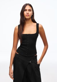 Pitkätummatukkainen nainen pukeutuneena istuvaan mustaan korsettityyliseen topiin ja korkeavyötäröisiin mustiin housuihin, poseeraa yhdellä kädellä taskussa.