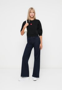 Hanorac negru cu un design în formă de inimă roșie, asortat cu blugi cu picioare largi de culoare albastru închis. Modelul ține o geantă albă, stând pe un fundal simplu.