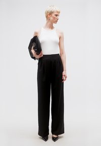 Ξανθό άτομο με λευκό αμάνικο τοπ και ψηλόμεσο μαύρο παντελόνι με φαρδιά ποδιά κρατάει μαύρο υφασμάτινο τσαντάκι, κοιτάζοντας προς τα πλάγια.