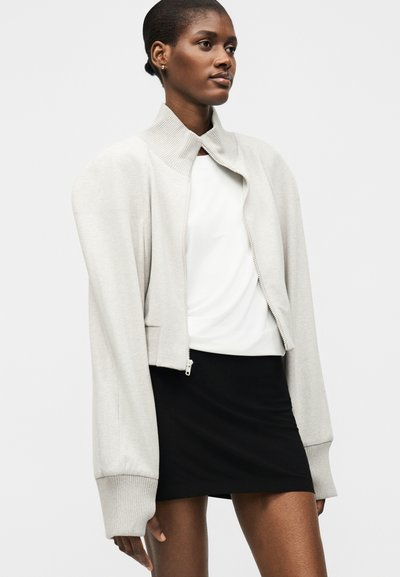 Γυναίκα που φοράει λευκό μπουφάν με ψηλό γιακά και φερμουάρ πάνω από λευκό τοπ και μαύρη φούστα, στέκεται μπροστά σε απλό φωτεινό φόντο.