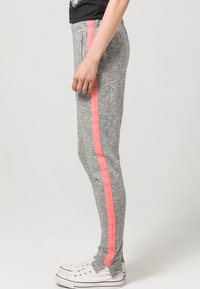 Graue Sweatpants mit schlanker Passform, ausgestattet mit einem pinken seitlichen Streifen und seitlichen Reißverschlusstaschen. Hergestellt aus weichem Material mit glatter Textur.