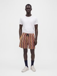 Homme debout portant un t-shirt blanc uni, un short rayé vertical multicolore, des chaussettes foncées et des baskets blanches sur un fond uni.
