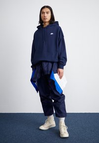 Ναυτικό φούτερ με κεντρική τσέπη και άσπρο λογότυπο, συνδυασμένο με παντελόνια με μπλε λεπτομέρειε�ς. Διαθέτει ανοιχτό μπεζ επιβλητικά αθλητικά παπούτσια.