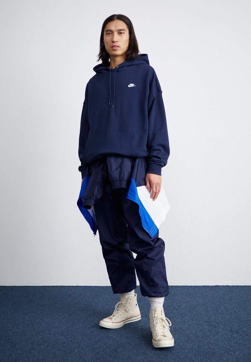 Granatowy hoodie z centralną kieszenią i białym logo, w parze z spodniami z niebieskimi akcentami. W zestawie jasnobeżowe sneakersy typu high-top.
