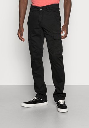 Mann trägt schwarze Cargohose mit Seitentaschen und schwarze Sneaker mit weißen Schnürsenkeln, steht auf grauem Boden vor einer weißen Wand.
