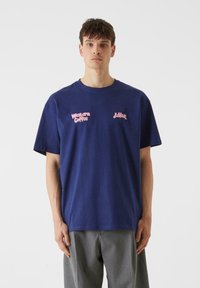 Mörkblå bomulls t-shirt med rosa grafisk text "Western Coffee" på vänster sida och "Jäkkar" på höger sida. Korta ärmar, avslappnad passform.