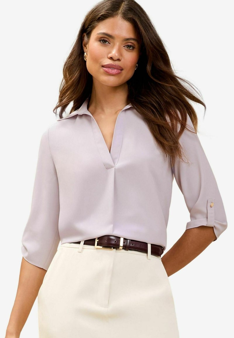 Blusa di un delicato lavanda con colletto, maniche a tre quarti, pattina frontale e una texture liscia, abbinata a una cintura marrone scuro e pantaloni crema.