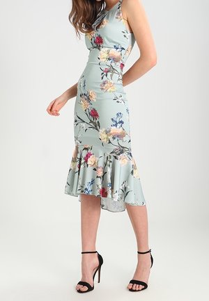 Kobieta w dopasowanej pastelowej sukience w kwiaty z falbaną u dołu i czarnych sandałach na wysokim obcasie z paskiem na kostce, stojąca na jednolitym tle.