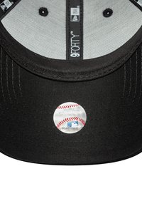 Melnā beisbola cepure ar gludu auduma virsmu, ar apļveida MLB logo uzlīmi iekšpusē un pelēku oderi, kas redzama cepures galā.