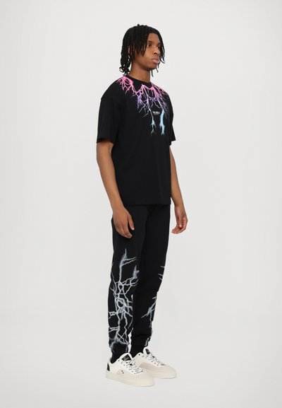 T-shirt nera con grafica di fulmini multicolore; abbinata a pantaloni neri con un design simile. Indossato con sneakers bianche.