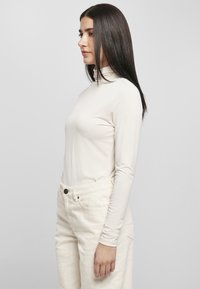 Maglia a maniche lunghe beige chiaro con colletto alto, realizzata in un tessuto liscio. Abbinata a pantaloni crema chiaro con bottone frontale e tasche.