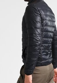 Zwarte pufferjas met een horizontaal doorgestikt patroon, een gladde glanzende textuur en een aansluitend ontwerp. Voorzien van een opstaande kraag en ritssluiting.