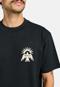 T-shirt di cotone nero con una grafica color crema che raffigura un'aquila con un design a raggio di sole e il testo "RVCA", maniche corte e scollatura rotonda.