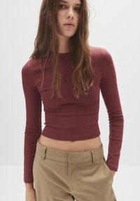 Langärmliges, burgunderrotes Cropped-Top aus dehnbarem Stoff, mit einem runden Halsausschnitt und einer taillierten Passform, kombiniert mit khakifarbenen Hosen.