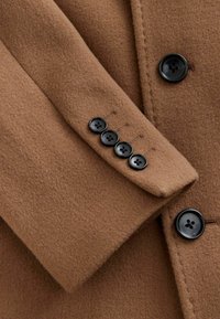 Details van een bruine wollen jas met een gestructureerde stof, een revers en vijf zwarte knopen op de manchetten met een ontwerp van vier gaten.