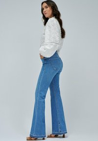 Jeans acampanados de denim en color azul, con una cintura alta, aberturas en los laterales del dobladillo y un corte ajustado. Combinados con una blusa blanca texturizada.