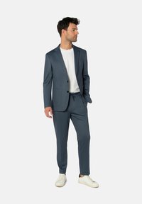 Maßgeschneiderter Herrenanzug aus dunkelblauem Stoff mit dezentem Muster, bestehend aus einem einreihigen Blazer und Kordelzughose. Kombiniert mit weißen Sneakern.