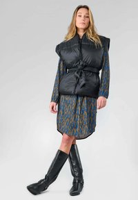 Gilet rembourré noir avec une ceinture à nouer, porté sur une robe à motifs bleu et marron. Le mannequin porte également des bottes noires en cuir montantes.