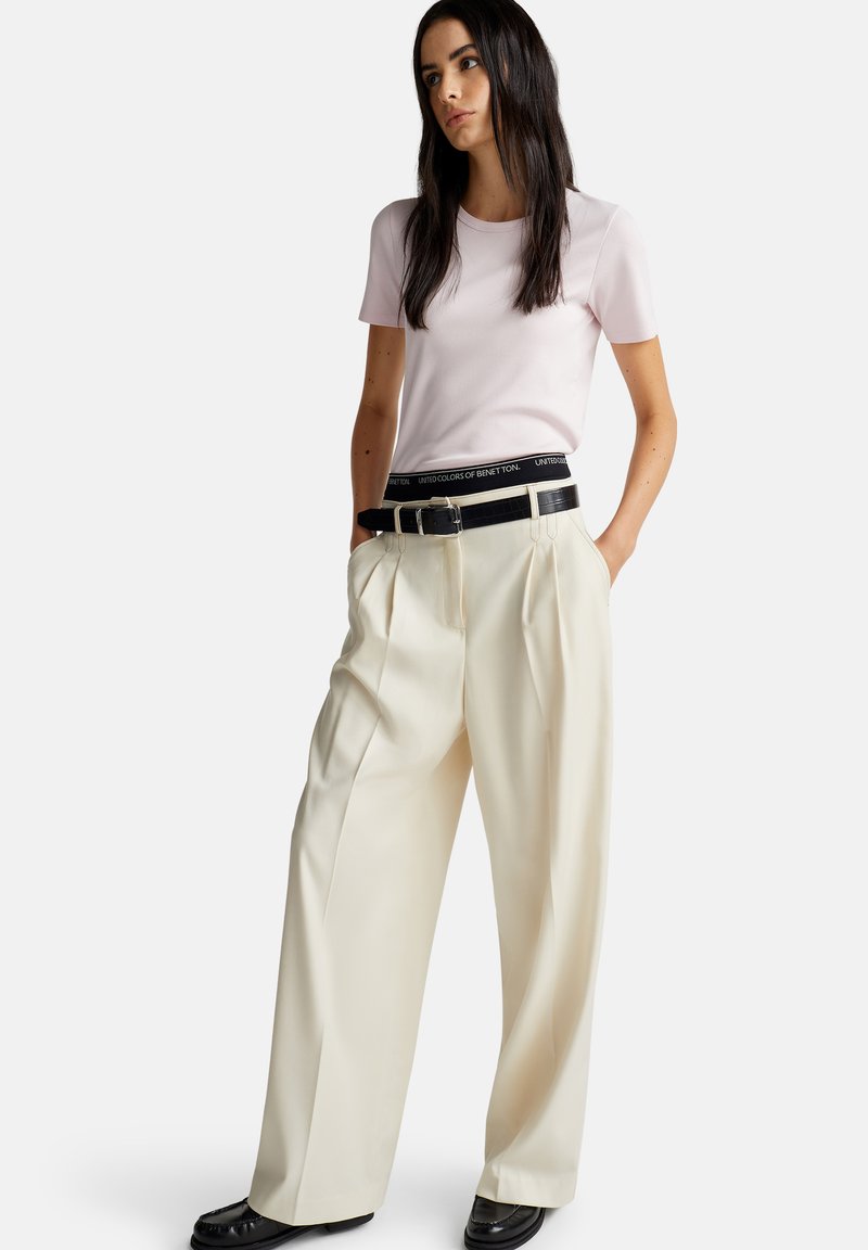 Světle růžové tričko s krátkým rukávem v kombinaci s krémovými plisovanými kalhotami s vysokým pasem a černým opaskem, který má lesklý povrch a detailní sponu.