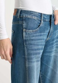 Personne portant un jean en denim bleu et un haut à manches longues blanc, avec une main reposant à côté et l'autre près de la poche.