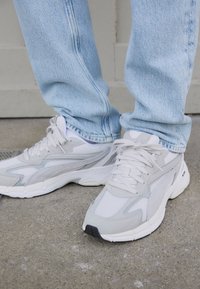 Light gray athletic shoes with mesh and suede details, featuring a textured sole and laces. Paired with light blue denim jeans.