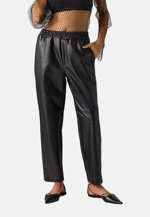 Pantalones de cuero sintético negros con cinturilla elástica, diseño de corte recto y textura suave. Llevados con unos zapatos planos negros de punta.