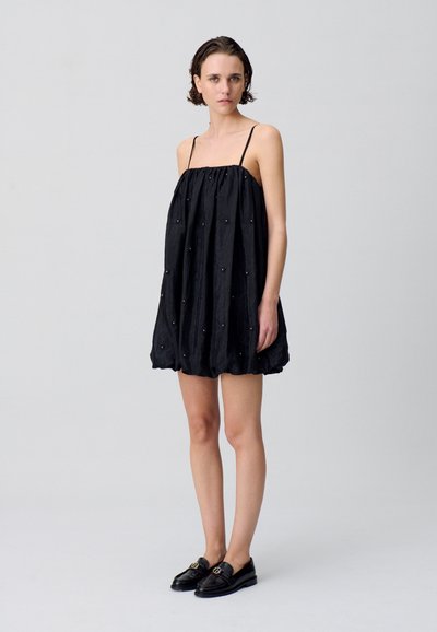 Vestido negro sin mangas con corte amplio y cuerpo fruncido. Presenta cuentas decorativas a lo largo del tejido. Se combina con mocasines negros.